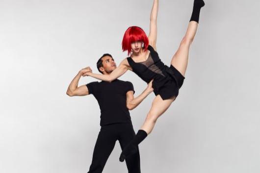 A man wearing all black supports another dancer wearing a red wig as does a tilt jump.